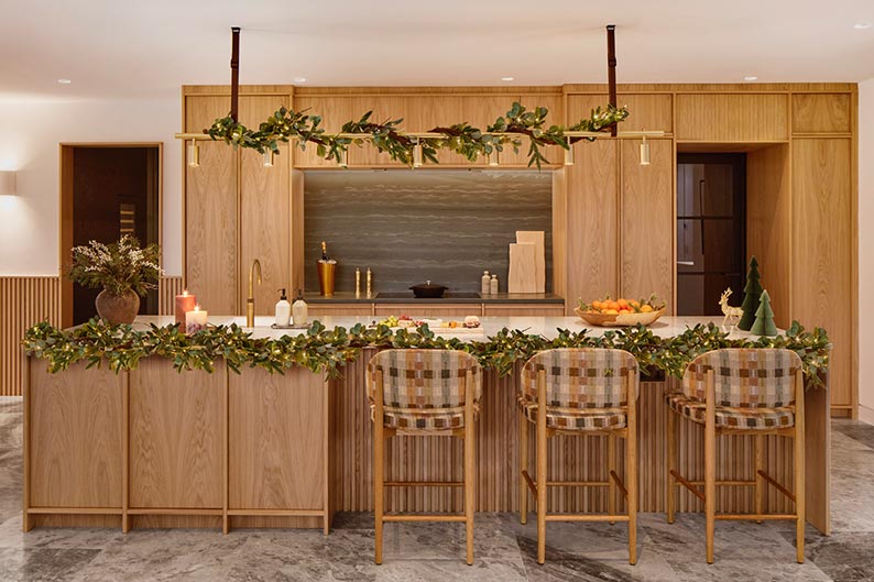 A large wooden kitchen with breakfast bar and three chairs. It's decorated with pine garlands along the worktop and lighting.